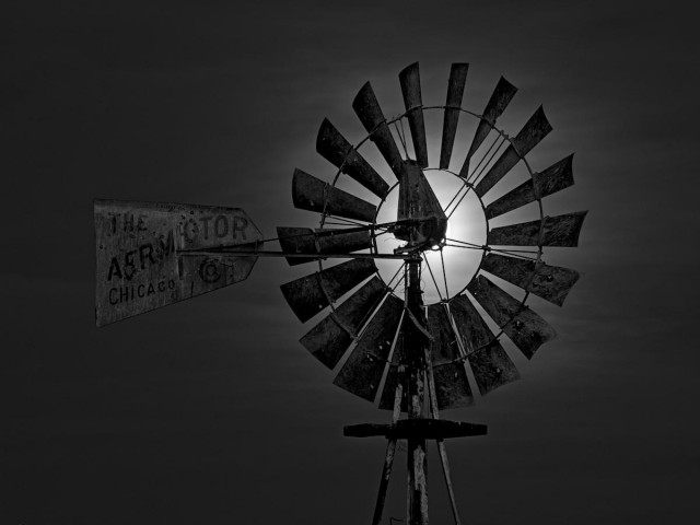 A black and white landscape photo of a windmill backlit by the Moon. The windmill is like a many bladed fan with a tail fin sticking out of the back. On the tail fin it says, "The Aermotor Chicago." The birght Moon is behind the hub of the fan and is thus lighting the area around the blades. otherwise, the sky is dark with a few streaks from a high thin layer of clouds.