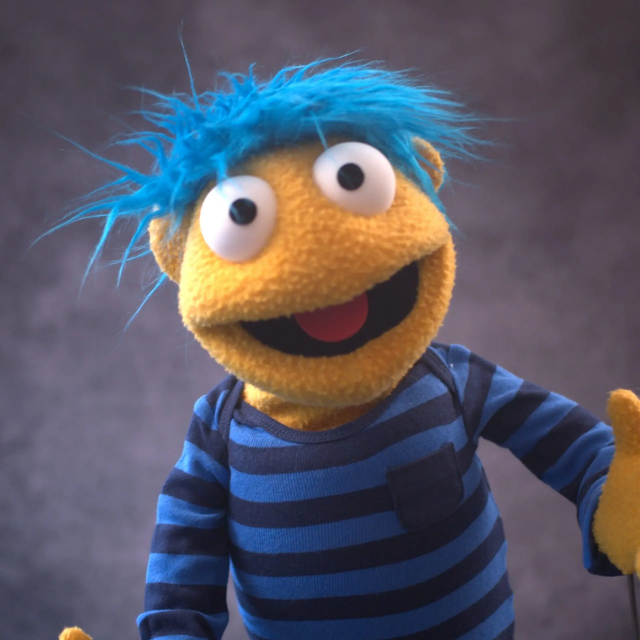 A yellow human puppet with blue hair in front of a photographic background 