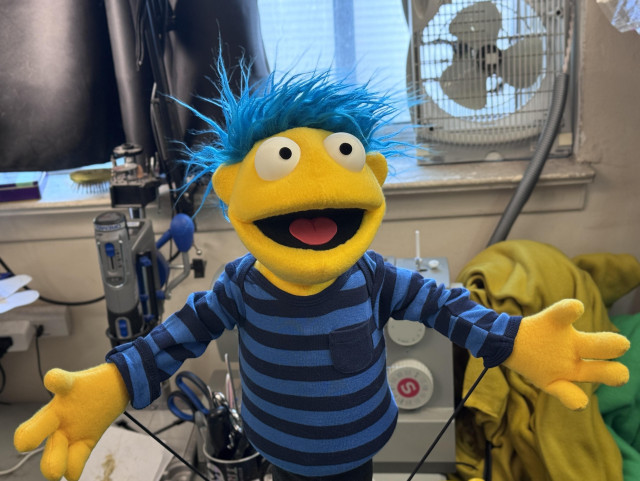 A yellow human puppet with blue hair on a work table