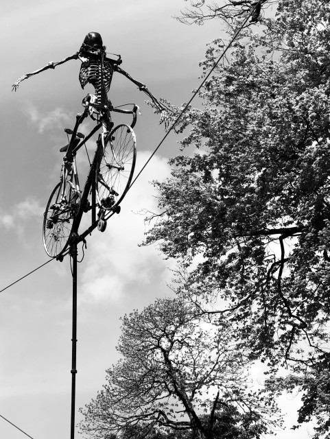 A skeletal figure on a vintage bicycle is riding a high-wire act, stretching out its arms for balance. The scene is set against a cloudy sky and surrounded by trees. The image is in black and white.