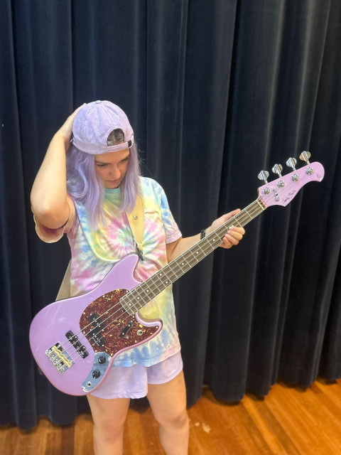 A girl with a lilac baseball cap, lilac hair, a pastel tie-dye shirt, lilac shorts, holding a lilac bass guitar. One hand is on the neck of the guitar, one on her head, she looking down, adjusting her hat. 

