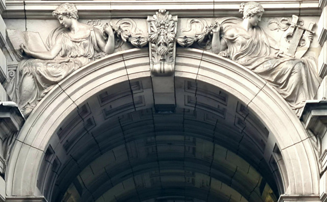 Two sculpted reclining female figures, nonchalantly holding up the ends of a central garland, while doing other activities. One is more focused in reading a book and the other on playing a lyre.