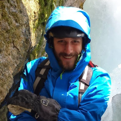 A dark haired, bearded man in a bright blue hooded ski jacket and black gloves, smiling.