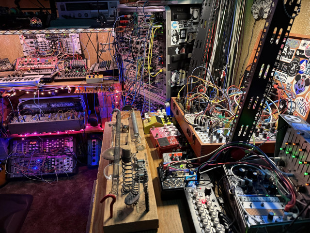 A busy music studio featuring various synthesizers, effects pedals, and modular equipment with colorful LED lights. Cables are everywhere. In the center on a table is a long experimental instrument.