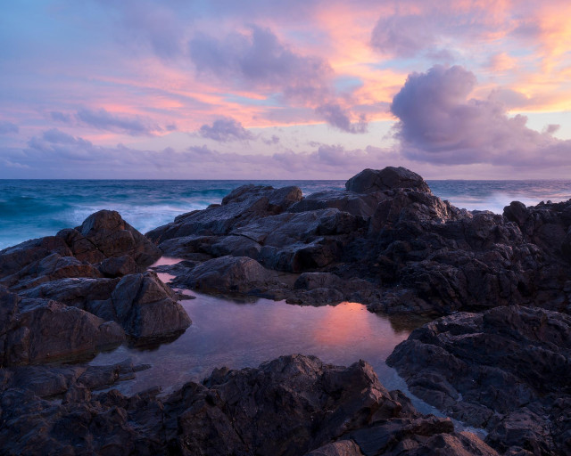sunset in pastel colours. A small puddle amongst the rocks at the top of the cliff. A stripe of the sea down below. Pastel coloured sky is reflecting in the puddle making it pink and slightly purple.