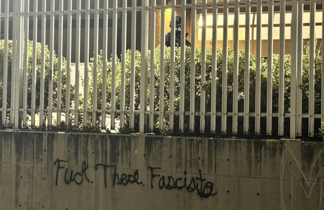 Tag on a wall on the side of the ice building says “fuck these fascists” and a fed is visible above the tag between the gate