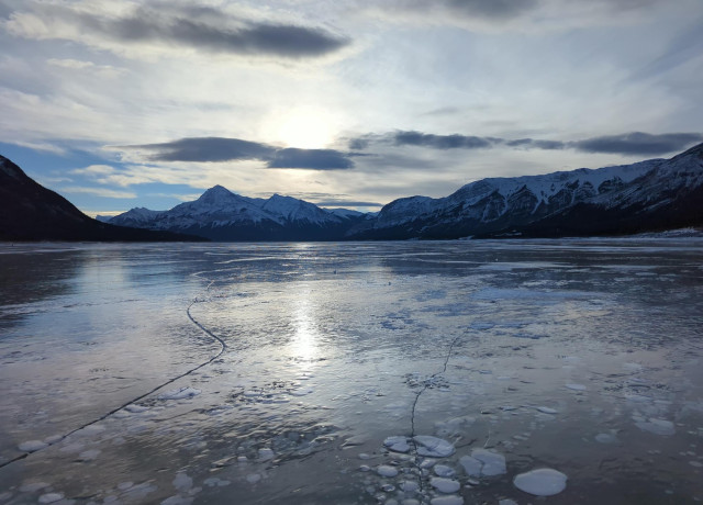 A view of a sunset while standing on Abraham Lake: a hydroelectric reservoir at the edge of the Rocky Mountains in Alberta, Canada. The image is split between the top half being a lightly snowy mountainous horizon and a partly cloudy sky, with gauzy clouds in-between fluffy ones. The sun is just off centre, is shining through these gauzy clouds, and is reflected in the lake's shiny surface

The bottom half of the image is the frozen lake's surface. It is quite shiny, having been polished by sun and wind, with only a few patches of snow in the distance. Several prominent surface cracks in the ice are visible, leading to the horizon. Embedded within the ice are both white and clear methane bubbles, giving a somewhat otherworldly feel. There is also a lightly bumpy but heavily polished surface texture to the ice, which is highlighted by the slanted light. 