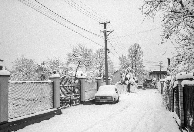 A quiet residential street covered in fresh snow. Low fences and gates line both sides of the narrow road, their tops thickly coated in white. A small car is parked along the fence, almost completely buried under snow, blending into the winter scene. Bare trees and bushes bend under the weight of snow, while power poles and overhead wires cut diagonally across the pale sky. The overall mood is still and subdued, with a sense of isolation and calm typical of an early winter morning