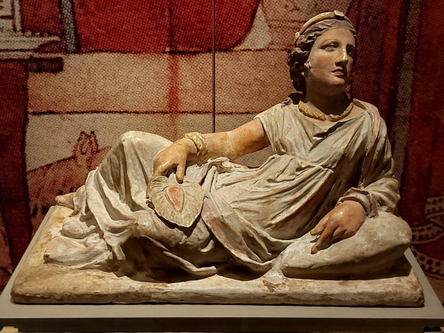 Painted Etruscan terracotta funerary urn lid  showing a reclining woman in banquet pose, holding a fan, wearing draped chiton, gold jewelry, on museum display. 