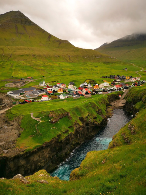 The image depicts a picturesque village nestled in a verdant valley, surrounded by lush green hills and mountains. In the foreground, a river flows through a narrow gorge, its waters a striking blue-green, contrasting with the surrounding greenery. The village is composed of colorful houses with red, green, and white roofs, some with distinct black accents, arranged along a winding road. A church with a white facade and a red roof stands prominently among the houses. The landscape is characterized by steep cliffs on either side of the river, with grassy slopes and patches of yellow wildflowers. The sky is overcast, with clouds partially obscuring the peaks of the mountains in the background, adding a dramatic effect to the scene.