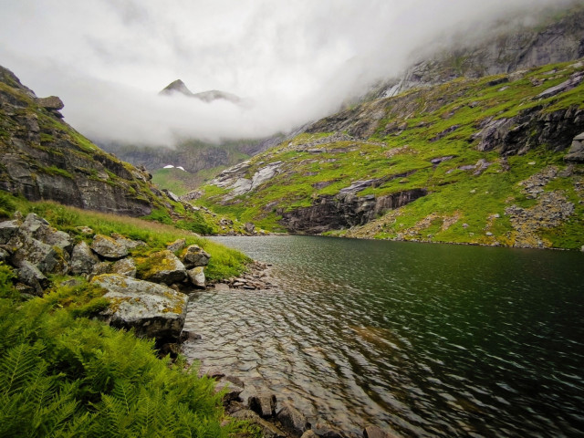 The image depicts a serene mountainous landscape with a calm lake at its center. The foreground features a rocky shoreline with large boulders covered in patches of moss and green ferns. The water of the lake is a deep green, reflecting the surrounding landscape. The midground shows steep, grass-covered slopes with rugged rock faces, and the background is dominated by towering mountains partially obscured by low-hanging clouds. The sky is overcast, contributing to the moody atmosphere of the scene. The overall color palette is dominated by greens, grays, and blues, with the mist adding a sense of depth and mystery to the image.