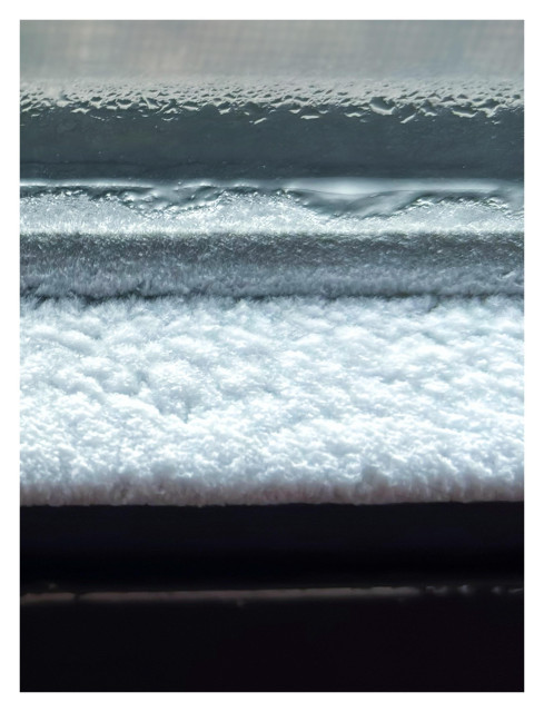A minimalistic image of horizontal bands of different widths and textures, in varying light and dark tones. It’s actually an extreme closeup of a layer of white frost on the lower lip of an interior window sash, with some drops of condensation on the glass above it and a blurred glimpse of the wire mesh screen on the outside.