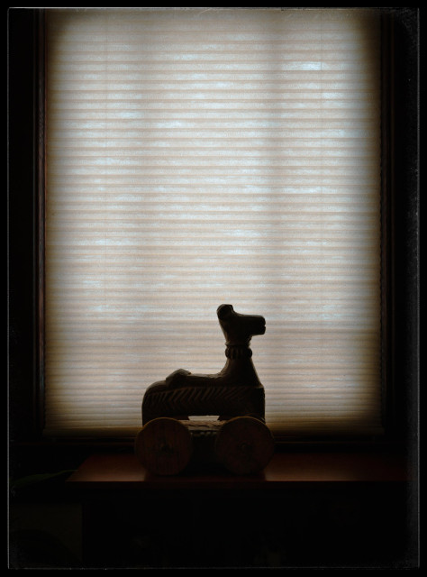 A silhouette of a dark stained, 10 inch, carved wooden toy sitting on a shelf in front of a cellular beige window shade. The simple carved horse has large, working wheels that rise up to just below the horse's belly.