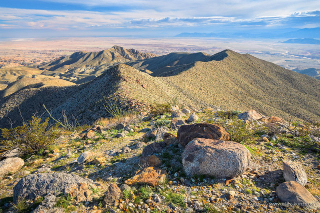 A color landscape photo with an elevated view across a vast desert landscape. Mid-afternoon light from the right creates nice depth and structure.  In the immediate foreground are big granitic rocks, the a ridge with rocks and small hardy shrubs. The ridge forms almost a mirrored S-shape, leading to a more distant and lower peak with eroded slopes. Beyond the peak lies a barren flat area, and all the way in the distance another mountain range. Some clouds hang in the sky over the distant mountains.