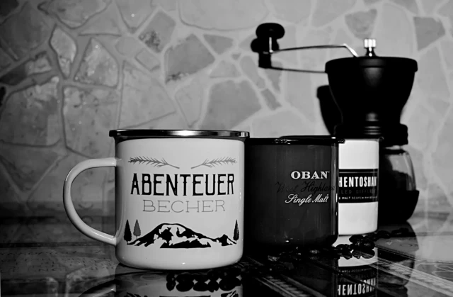 Drei Emaille-Tassen stehen in einer Reihe auf einer Glasoberfläche. Die vordere Tasse zeigt ein Bergmotiv, überschrieben mit dem Wort Abenteuer. Die mittlere ist eine Tasse aus der Oban Whisky-Destillerie, die hintere zeigt das Label der Whisky-Destillerie Auchentoshan. Ganz hinten steht eine handbetriebene Kaffeemühle.