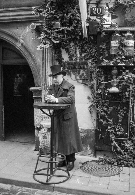A black-and-white photograph taken on an old street in Lviv.
An elderly man in a long dark overcoat stands quietly by a tall, round metal table on the sidewalk. On his head he wears a top hat, which gives him a dignified, slightly old-fashioned appearance. His hands are folded, and his posture is calm and contemplative, as if he is waiting or absorbed in thought.

Behind him, the weathered façade of an old building is partially covered with ivy, revealing layers of age and texture. An arched doorway opens into shadow, while vintage lamps, bottles, and exposed pipes create a subtly surreal, timeworn atmosphere. The street and architectural details evoke a strong sense of history, lending the scene a nostalgic, almost cinematic quality—as if the moment belongs to another era.