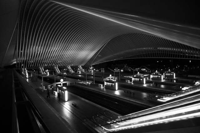 Schwarz-weiß Foto vom Bahnhof in Lüttich abends in der Dunkelheit. Der Bahnhof ist architektonisch sehr interessant, er hat ein hohes geschwungenes Dach. 