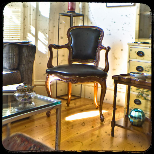 A through-the-viewfinder TtV view of a Victorian living room, with hardwood floors, glass and wooden side tables, an antique chair, tall antique chest, treasured glassware, and a bit of an oriental rug. There is light shining into the room from shuttered windows that face the street.

The square cropped photo is full of specs and dust, and is heavily distorted around the edges. 

Photographed using a modern Pentax DSLR that is peering down into an old Kodak Duaflex twin lens reflex camera.