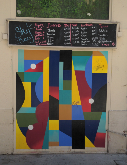 Sur une large porte en bois, sous le menu du "Sky bar", une grande fresque rectangulaire, en formes géométriques imbriquées, jaunes, bleues, noires, rouges et vertes.