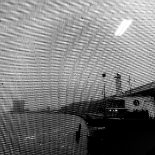 Black and white square photo looking through textured glass out across the water behind Amsterdam's Central Station (part of which is visible on the right of the frame). The background sky is overcast. Near the top right of the frame is a reflection of a light on the window glass.