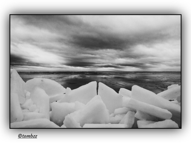 This black-and-white photograph captures a frozen bay shoreline, where jagged ice ridges known as pressure ridges or hummocks have piled up near the edge. These formations arise when shifting ice sheets collide and crack under wind stress or temperature changes, forcing slabs of ice to thrust upward. In the image, they form a chaotic, serrated wall, like a wave frozen mid-motion. Above the bay, heavy, layered clouds hang low and dense, as if the sky itself were about to break. The ice stretches unbroken to the horizon, and the scene radiates cold, tension, and a stark, arctic stillness.