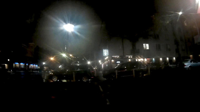 ein bei Dunkelheit mit einer am Fahrrad montierten Dashcam aufgenommenes Bild. Es ist mittig unten eine weisse, gestrichelte Linie zu erkennen die zur Bildmitte hin auf zwei Autos zu läuft, die quer zu einen Fuss- und Radweg abgestellt sind, so das nur eine Lücke bleibt durch die sich ein Fussgänger zwängen kann. 