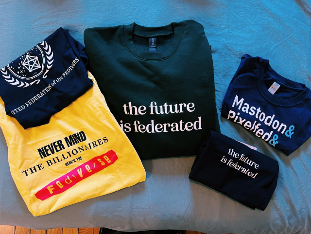 a photo of several t-shirts and sweatshirts folded on a teal bedspread. Top left: navy blue t-shirt that reads "united federation of the fediverse" under it a yellow t-shirt that reads "never mind the billionaires here's the fediverse". Center: a dark green sweatshirt that reads "the future is federated". Right: a blue t-shirt that reads "Mastodon& Pixelfed&" and below it a black t-shirt with "the future is federated" written in white