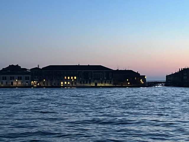 Colour photo, taken just after sunset, looking out over a wide canal at the waterfront of the island beyond. The two-storey building in the centre of the photo is long and low, and mainly in darkness, apart from a cluster of windows, three on the ground floor and four directly above, which are lit from inside and glow a warm inviting yellow. To the right of the building is a gap in the skyline for a barely-visible bridge, and then the corner of a bark block of a building on the right edge of the frame. The expanse of water in front is steel-blue and gently rippled, and the sky above is in the cold pastel shades of gloaming, rising from red through purple to blue. The image feels somber but tranquil. 