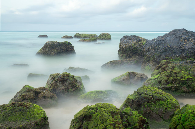 seascape with mossy rocks, long exposure smoothed water and cloudy sky. The color palette is green and cyan and slightly sad