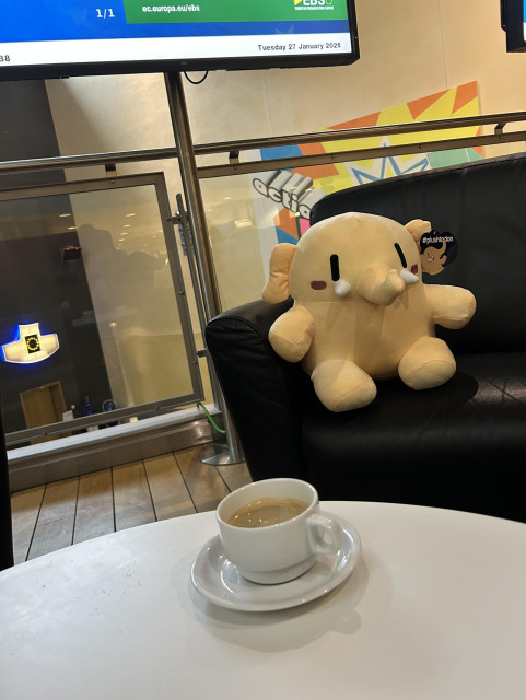 In the foreground is a cup of coffee on a table, then in the background the mastodon plushie sitting on a black leather sofa. 