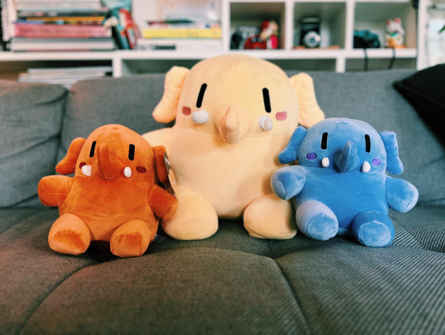 A photo showing my Plushtodon family on the couch. Mini orange plushie on the left, big beige Plushtodon in the center and mini blue Plushtodon on the right 
