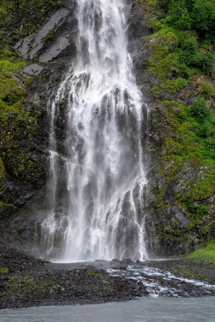 A vertical image of Bridal Veil Falls in Keystone Canyon near Valdez, Alaska. A tall, narrow waterfall descends down a steep, rocky cliff covered in dense green vegetation. At the base of the falls, water flows into a gray, fast-moving river. The rock walls rise sharply on both sides of the frame.