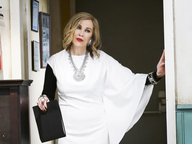Catherine O'Cara as Moira Rose wearing a long white dress with one long sleeve and one short, revealing a black top underneath it, some extravagant jewellery, and red lipstick. 