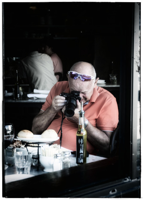 A picture, taken from the street, of a man seated inside a restaurant, at a cafe table. He is wearing a peach colored polo shirt and has cycling sunglasses flipped up on top of his head. Most importantly, he has a Nikon DSLR in his hand and his attention is focused on capturing an Instagram-worthy picture of the food on his plate.

In 2014, taking food photos was a requirement, not an option.