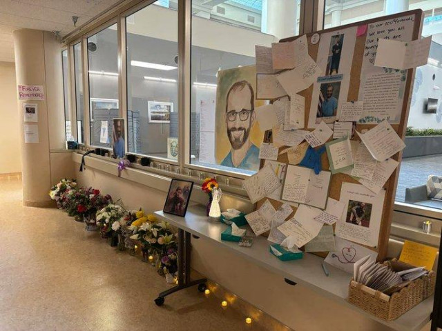 A picture of a windowed corridor inside the hospital, with flowers and candlces lining the floor.  Against the window, a large noticeboard with messages of condolence and memory pinned to it sits alongside a painting of Alex.