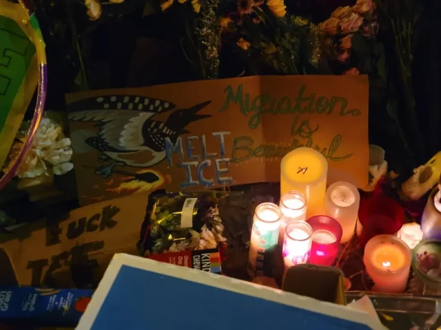 A picture of flowers, candles and messages at Alex's shrine.  At the centre is a large cardboard sign with a picture of the loon (Minnesota's state bird) flying, and the words MIGRATION IS BEAUTIFUL, MELT ICE.
