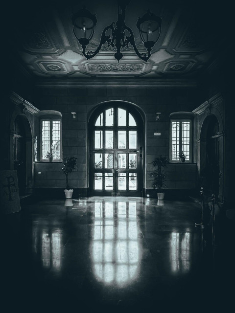 Eine Eingangshalle mit einer großen gewölbten Tür am Ende. Der Raum verfügt über eine dekorative Decke, Kronleuchter und Topfpflanzen. Reflexionen sind auf dem polierten Boden sichtbar

An entrance hall with a large arched doorway at the end. The space features a decorative ceiling, chandeliers, and potted plants. Reflections are visible on the polished floor.