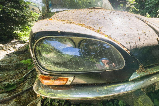 Close-up of a dirty, weathered car headlight with leaves and debris on the hood; a person is reflected in the glass.