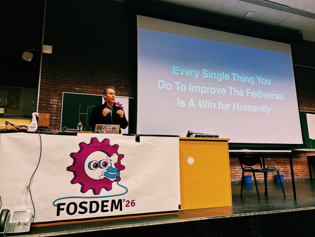 A photo showing Ben Pate on stage at FOSDEM in the Social Web devroom. He’s standing behind a table that has the FOSDEM ‘26 logo and next to him there is a large screen showcasing a slide that reads “Every Single Thing You Do To Improve the Fediverse Is A Win for Humanity”
