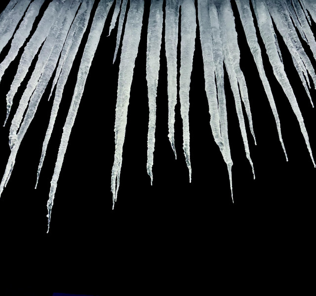 Photo of icicles at night. The background is completely dark and the icicles are lit up by a light behind the camera. 

I took this photo in February, 2025 in Stowe, Vermont. The icicles were hanging down from the edge of the roof of a local restaurant. The icicles were quite large— about 3 feet long.
