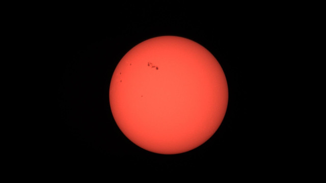 A bright, reddish-orange sun with small dark spots on its surface, set against a pitch-black background.