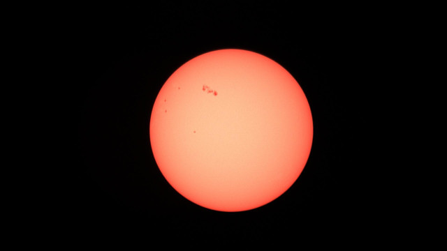 A bright, pinkish-orange sun with some dark sunspots against a black sky.