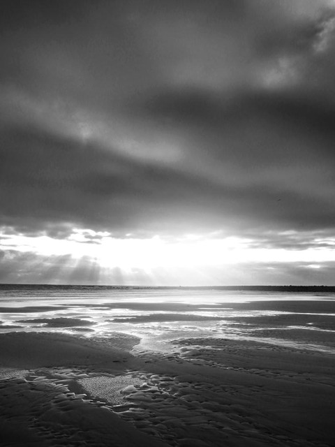 Atmospheric black and white photo of sunlight reflected on a beach at low tide, with dark clouds overhead.