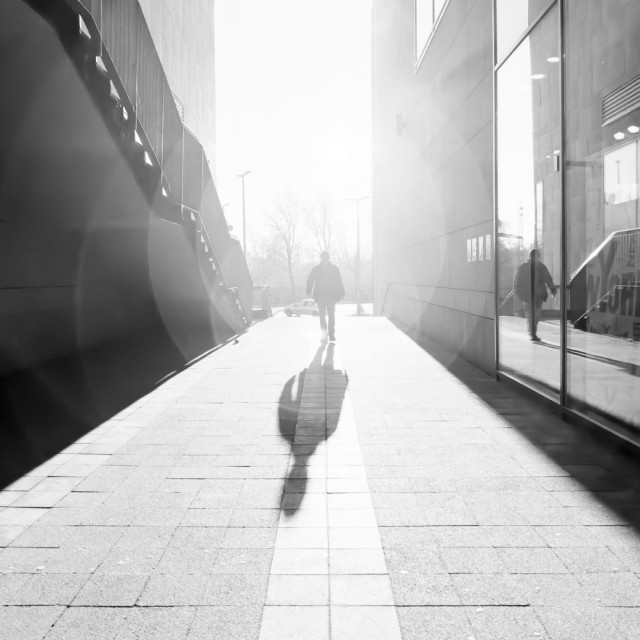 Ein Gang zwischen einer Treppe und einem Schaufenster. Gegen die Sonne eine Entgegenkommender, der sich auch im Schaufenster spiegelt. Quadratisch, s/w.
Hell.