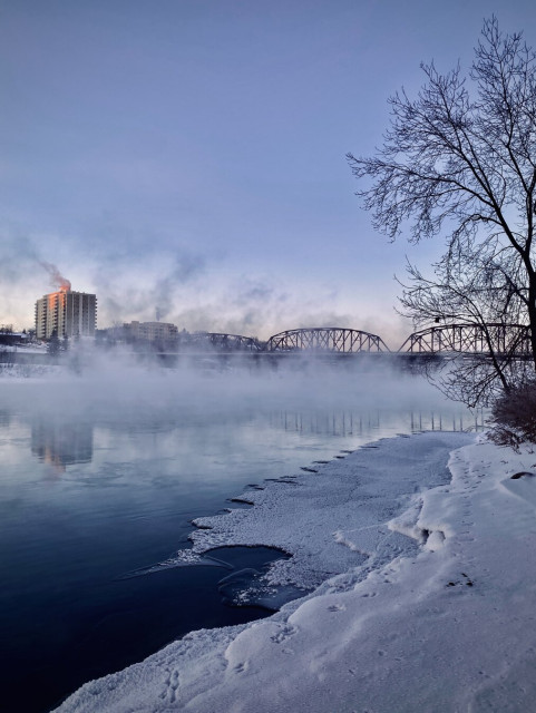 View from the icy edge of a river, mist rising and partially obscuring the opposite shore. A metal bridge in the background and a tall building highlighted by the pink light of the sunrise is reflected in the water. The sky is a clear blue.