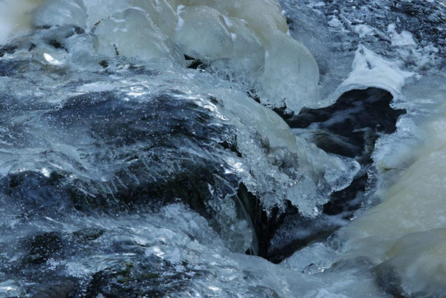 Part of a forest brook that is almost totally covered up in ice, some of it is thick, coloured by iron in a pale yellow tone, and some of it is thin and reminds of glass where you can see the water running under it. A bit to the right of center a small part is still open and you can see the dark water running down the brook. 