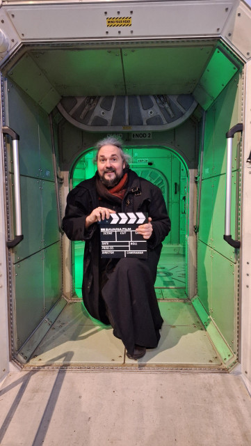 Ein lächelnder Herr mit etwas längeren Haaren, der eine Filmklappe in Händen hält. Er trägt einen schwarzen Mantel und kniet in einem grün beleuchteten Durchgang, der ein wenig nach Raumstation aussieht.