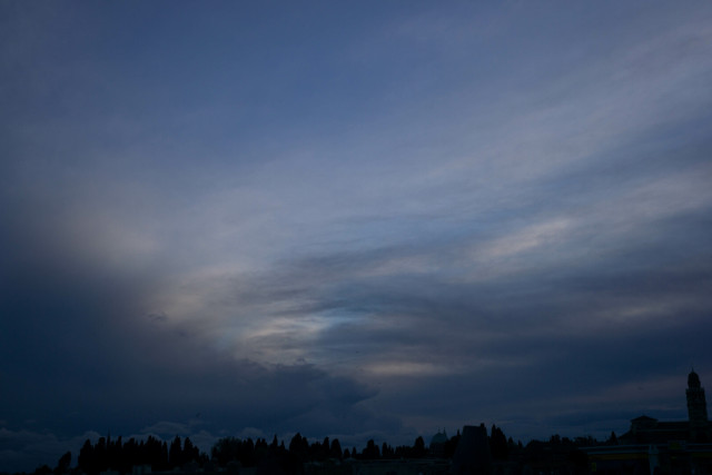 Photo looking up at a cloud-streaked sky, it gets dark towards the lower-left and lower-right corners. Across the very bottom are silhouettes of low-rise buildings, including at least one dome and one domed tower.