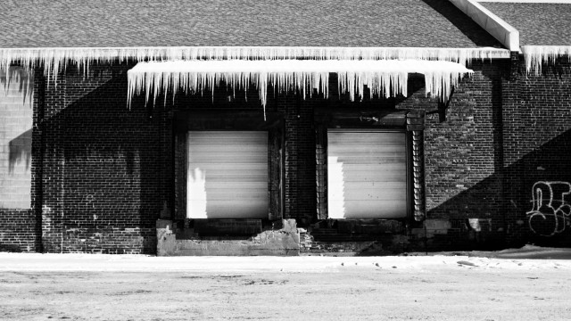 Large icicles hang from a building and from a overhang protecting two large loading doors. This is a black and white image.