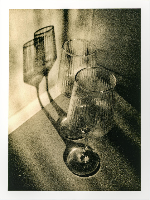 A moody lith print depicting two empty drinking glasses placed close together on a flat surface near a wall. Their translucent bodies catch an angled light, casting elongated, slightly distorted shadows that echo their shapes. The grainy, high-contrast texture typical of lith printing breaks the image into speckles and warm, uneven tones, blurring the boundary between object and shadow. The scene feels quiet and contemplative, as if the glasses are traces of a recent presence—something just finished, or already fading.